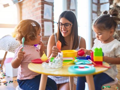 Young beautiful teacher and toddlers playing meals using plastic food and cutlery toy at kindergarten